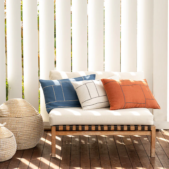 A stylish outdoor bench with three decorative pillows in blue, white, and terracotta, featuring geometric stitching. Woven ottomans rest nearby, set against vertical white paneling and a wooden deck, showcasing a modern and cozy decor collection.