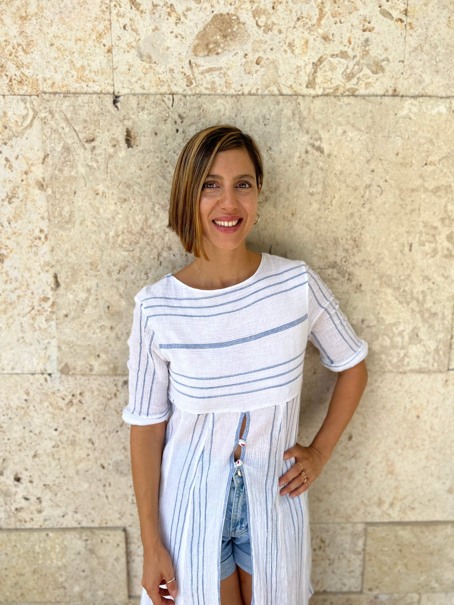 Home Tailor founder, Gizem Acar, stands in front of a textured beige stone wall. She has a radiant smile and shoulder-length hair, and is wearing a casual, stylish white tunic with thin blue stripes, a front tie detail, and paired with denim shorts. Her relaxed pose and warm expression evoke a sense of effortless summer fashion and tropical charm.